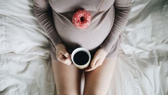 vrouw die met een kopje koffie en een donut op haar zwangere buik zit te genieten zwanger beter niet eten