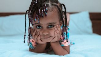 Meisje dat op bed ligt met haar hoofd in haar handen en bijdehand de camera in kijkt