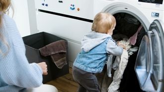 Kindje die de was in de wasmachine doet, terwijl haar ouders deze moeten schoonmaken omdat hij stinkt