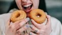 vrouw houdt twee donuts voor haar mond, alsof ze veel gaat eten: een van de beschamende dingen die we doen