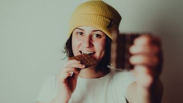 Vrouw met gele muts die chocolade eet en een stukje aanbiedt aan de camera