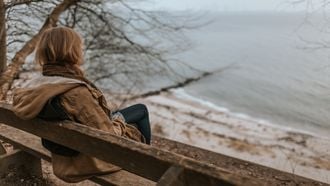 Vrouw zit op een bankje en kijkt uit over zee, ze vraagt zich af of ze overspannen is of een burn-out heeft