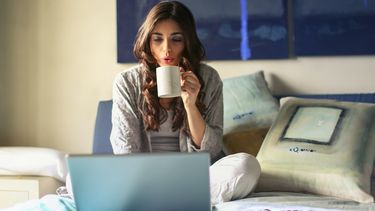 Vrouw zit bij thuiskomst op bed met laptop en kop thee