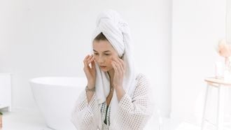 Vrouw in witte badjas en handdoek op haar hoofd brengt zalf aan op haar gezicht. Skincare in de koelkast