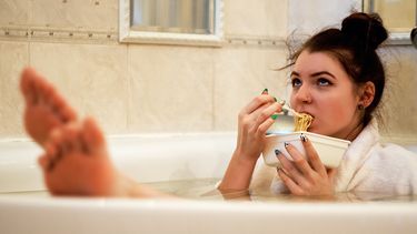 Vrouw die alleen thuis is en in haar badjas in bad zit met een bord pasta